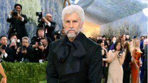 Bearded man with white hair in a black suit and oversized bow tie poses on a red carpet as photographers crowd behind him.