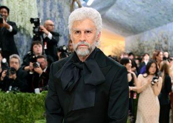 Bearded man with white hair in a black suit and oversized bow tie poses on a red carpet as photographers crowd behind him.