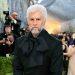 Bearded man with white hair in a black suit and oversized bow tie poses on a red carpet as photographers crowd behind him.
