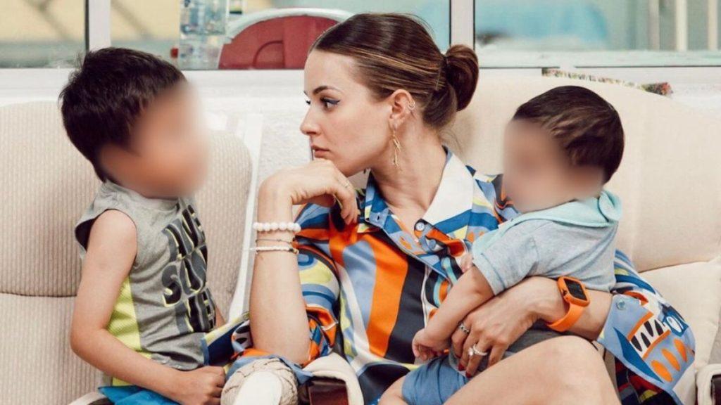 Mother sitting on a couch with two young children, one on her lap and the other beside her; both children's faces are blurred for privacy.