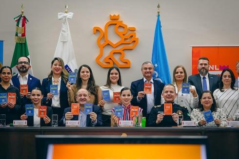 Group of professionals in a conference room posing behind a long table, holding orange and blue certificates, with a crest on the wall and flags in the background.