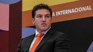 Man in a dark suit and orange tie seated on stage at an international conference, looking to his left.