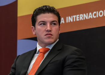 Man in a dark suit and orange tie seated on stage at an international conference, looking to his left.