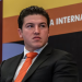 Samuel García señalado por triangular más de 200 millones de pesos a una de sus empresas - Minuto Cero Man in a dark suit and orange tie seated on stage at an international conference, looking to his left.
