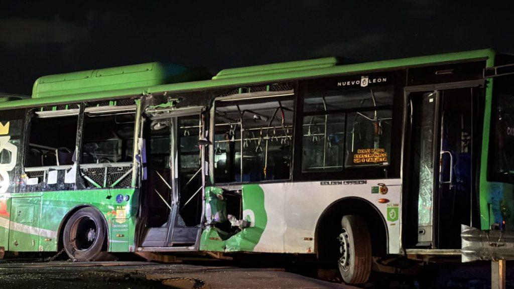 Green and white city bus with extensive front-left damage and open doors at night, inside showing dangling handrails and seats, suggesting a traffic accident.