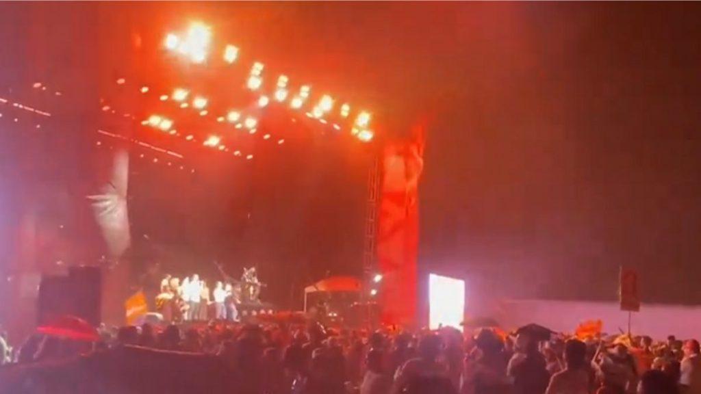 Crowd at a nighttime outdoor concert watching a stage bathed in red and orange lights with performers on the left.