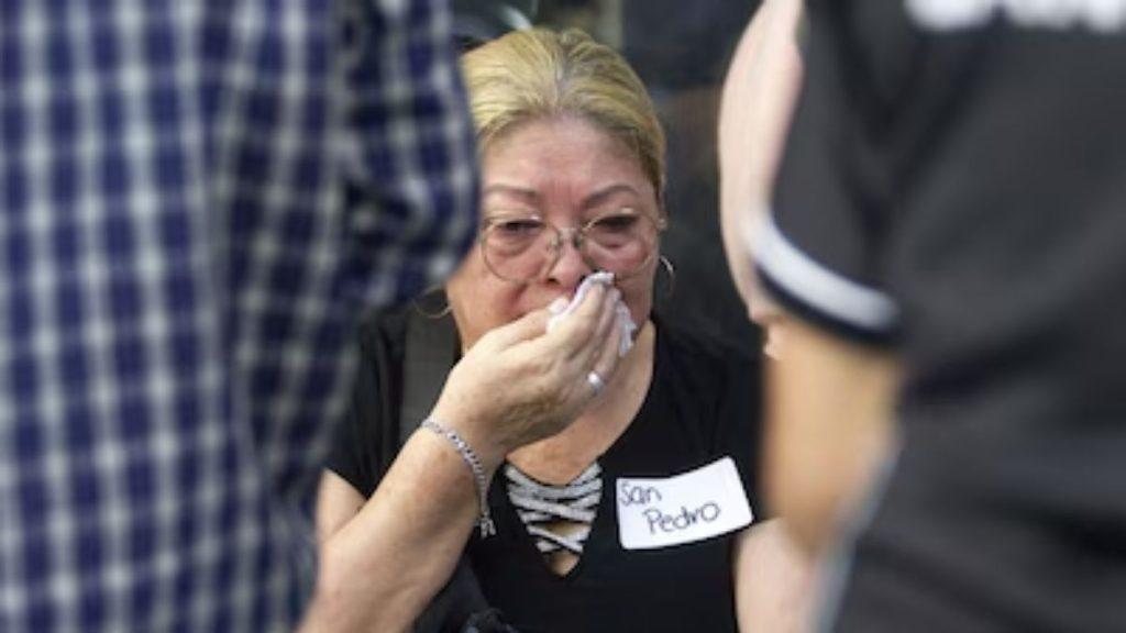 Older woman with glasses wipes her nose with a tissue at a crowded gathering, wearing a black top and a name tag on her chest.