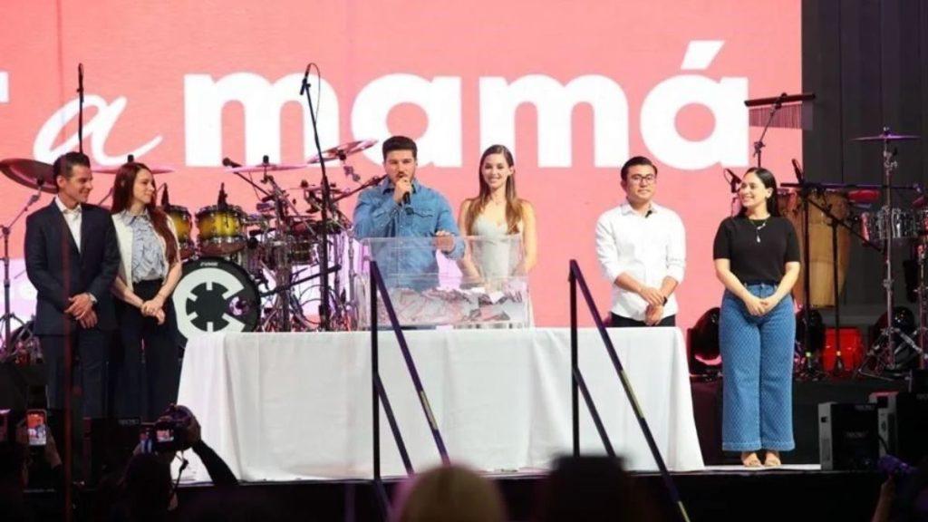 Six people on a stage behind a white table, a man speaks into a microphone with a pink backdrop.