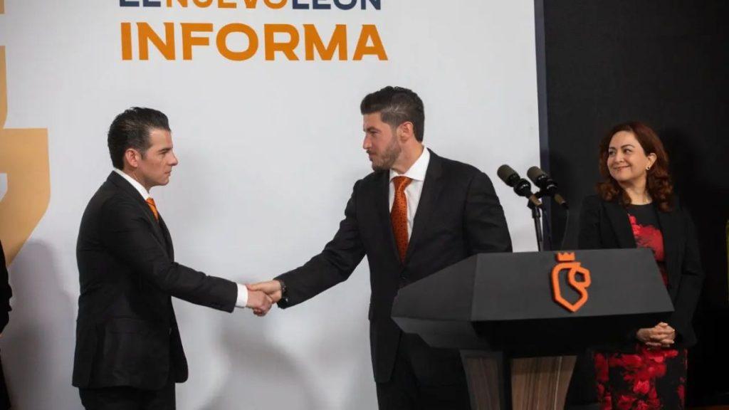 Two men in dark suits shake hands on a stage beside a black podium with an orange emblem, while a smiling woman stands nearby.