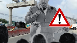 Speaker addressing a crowd at a road construction site with a large warning triangle overlay indicating caution.