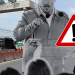 Speaker addressing a crowd at a road construction site with a large warning triangle overlay indicating caution.